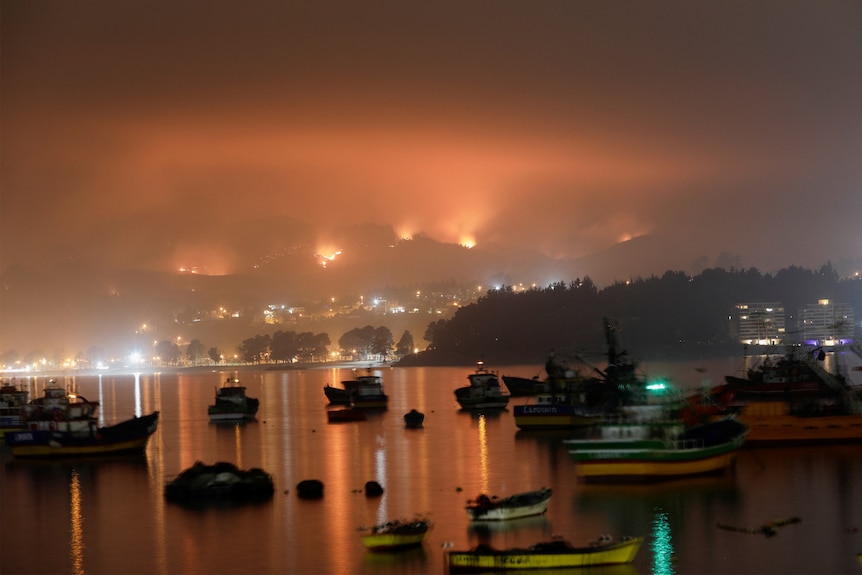 A harbour with several boats in it with a red smokey haze over it with flames in the background