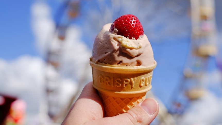 A hand holding an ice cream cone topped with a straberry, with a ferris wheel in the background. 