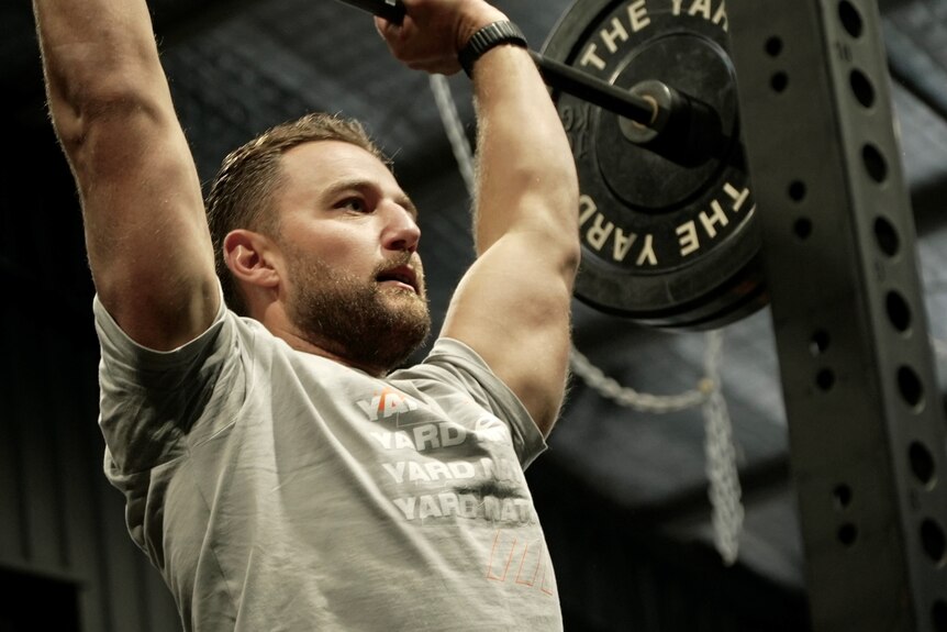 Daniel Bova, wearing a grey T-shirt, with short brown hair and a neat beard, pumps iron in the gym.