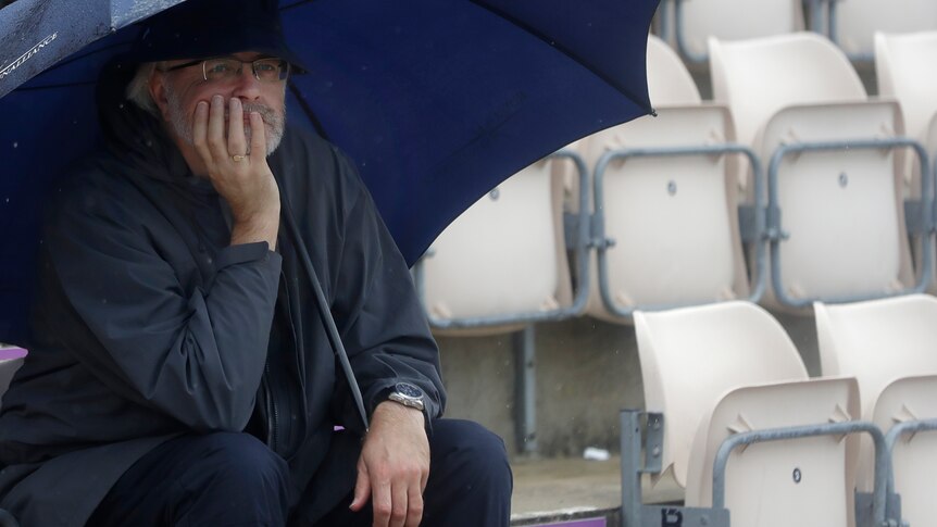 A man wearing a blue rain jacket shelters under an umbrella leaning on his hand with empty white seats next to him