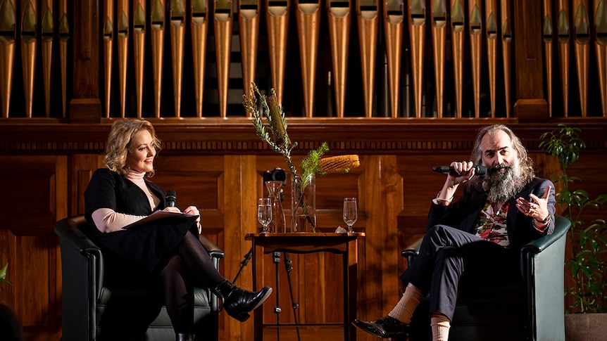 Zan Rowe and Warren Ellis sit in front of massive pipe organ at Hobart Town Hall. Warren speaks into a microphone.