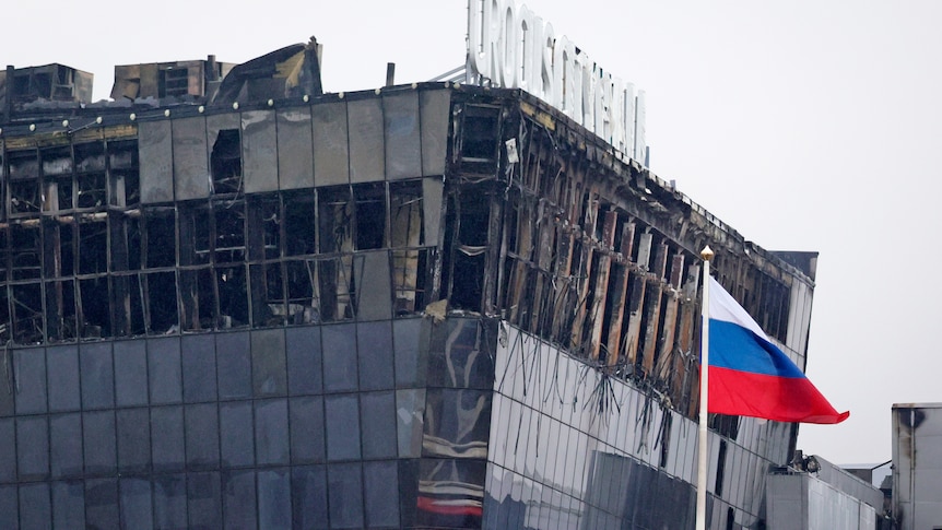 A large building with burn marks and shattered windows sits against a grey sky with a russian flag flying in front of it