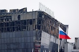 A large building with burn marks and shattered windows sits against a grey sky with a russian flag flying in front of it