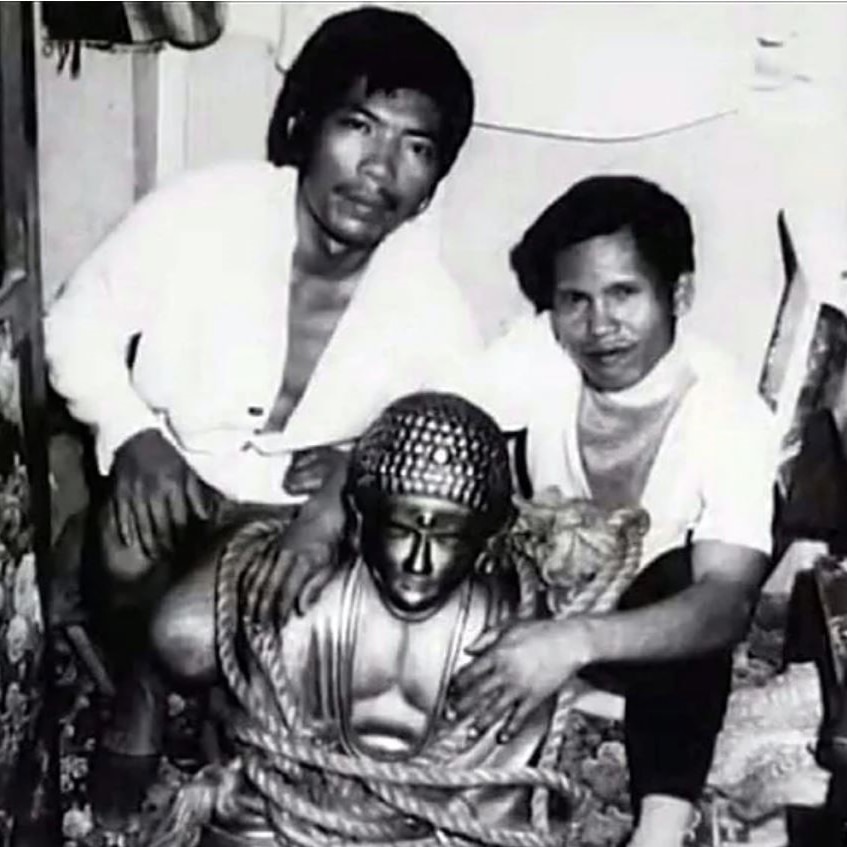 A black and white photo of two men crouching next to a shiny buddha statue wrapped in a rope 