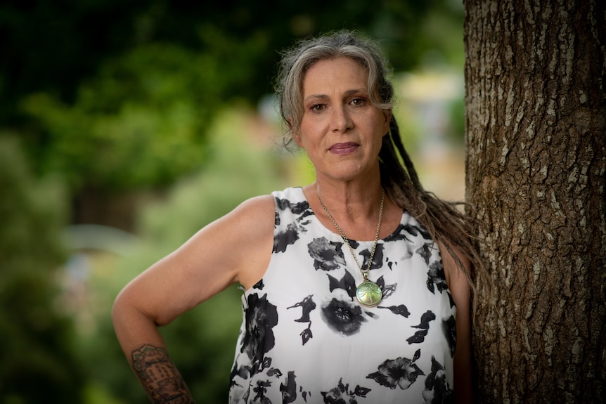 A woman stands next to a tree trunk with one hand on her hip, looking directly into the camera