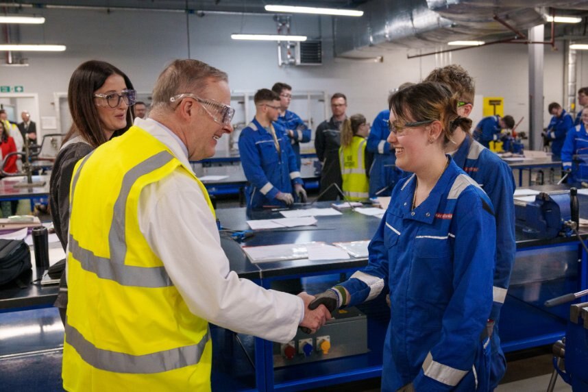 Albanese, in high vis, shakes the hand of a young woman in a blue coverall in a workshop