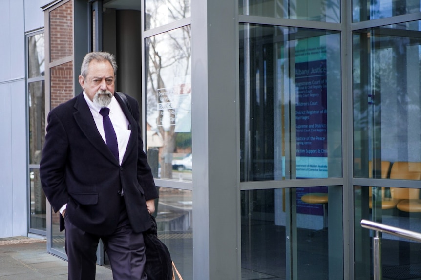 A grey-haired man with a short beard and wearing a black suit jacket walks away from a glass-fronted building.