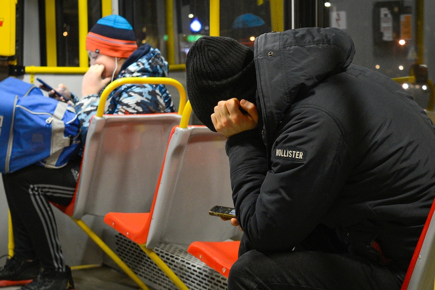 A man in a beanie sits on a bus, holding his head and looking at his phone.