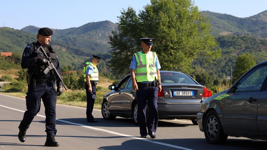 Police are pictured walking down a road with a soldier holding a gun. Cars have been pulled over.