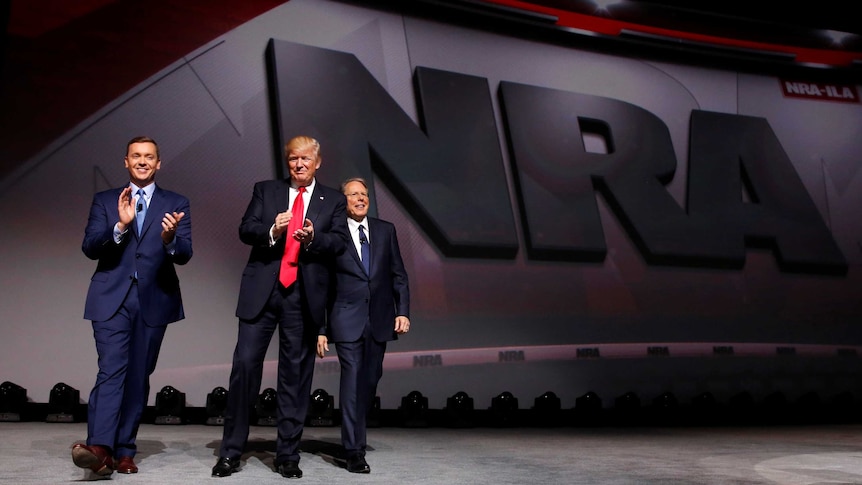 Three men in suits smile and clap in front of a huge NRA logo.