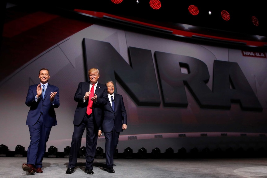 Three men in suits smile and clap in front of a huge NRA logo.