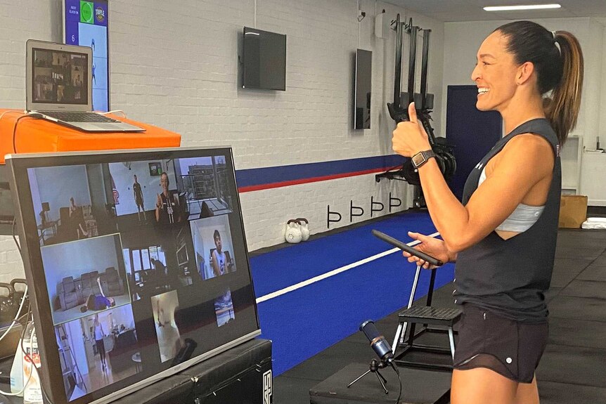 A woman hosts a gym working in front of a big Zoom video conference monitor.