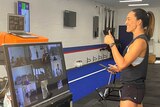 A woman hosts a gym working in front of a big Zoom video conference monitor.