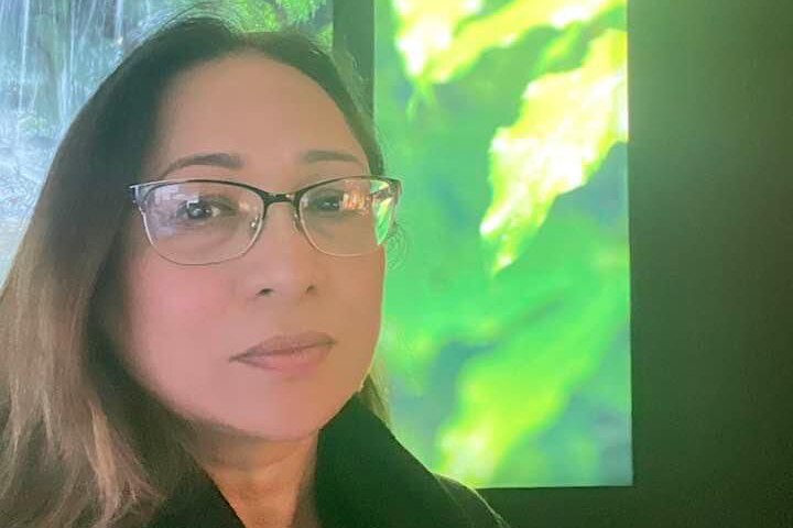 Woman with glasses is wearing black top, there's a window in the background showing green leaves.