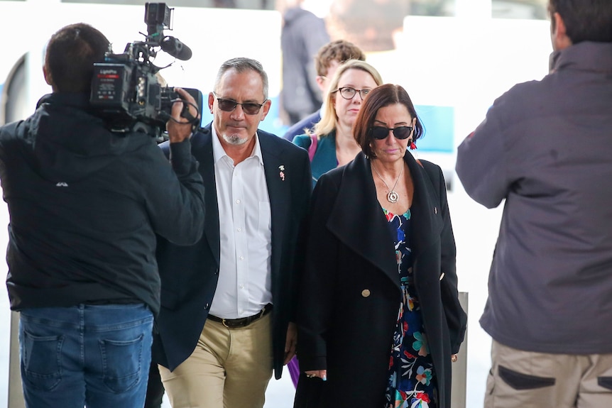 Sue and Lloyd Clarke walk through a crowd of camera operators.