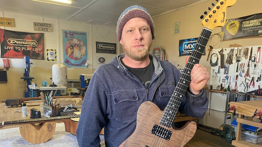 A man wearing a beanie holding an electric guitar in a guitar making workshop.