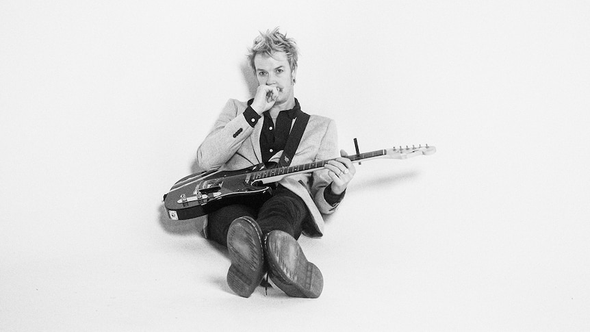 Black and white photo of Chris Cheney sitting on the floor with his legs stretched out, holding a guitar