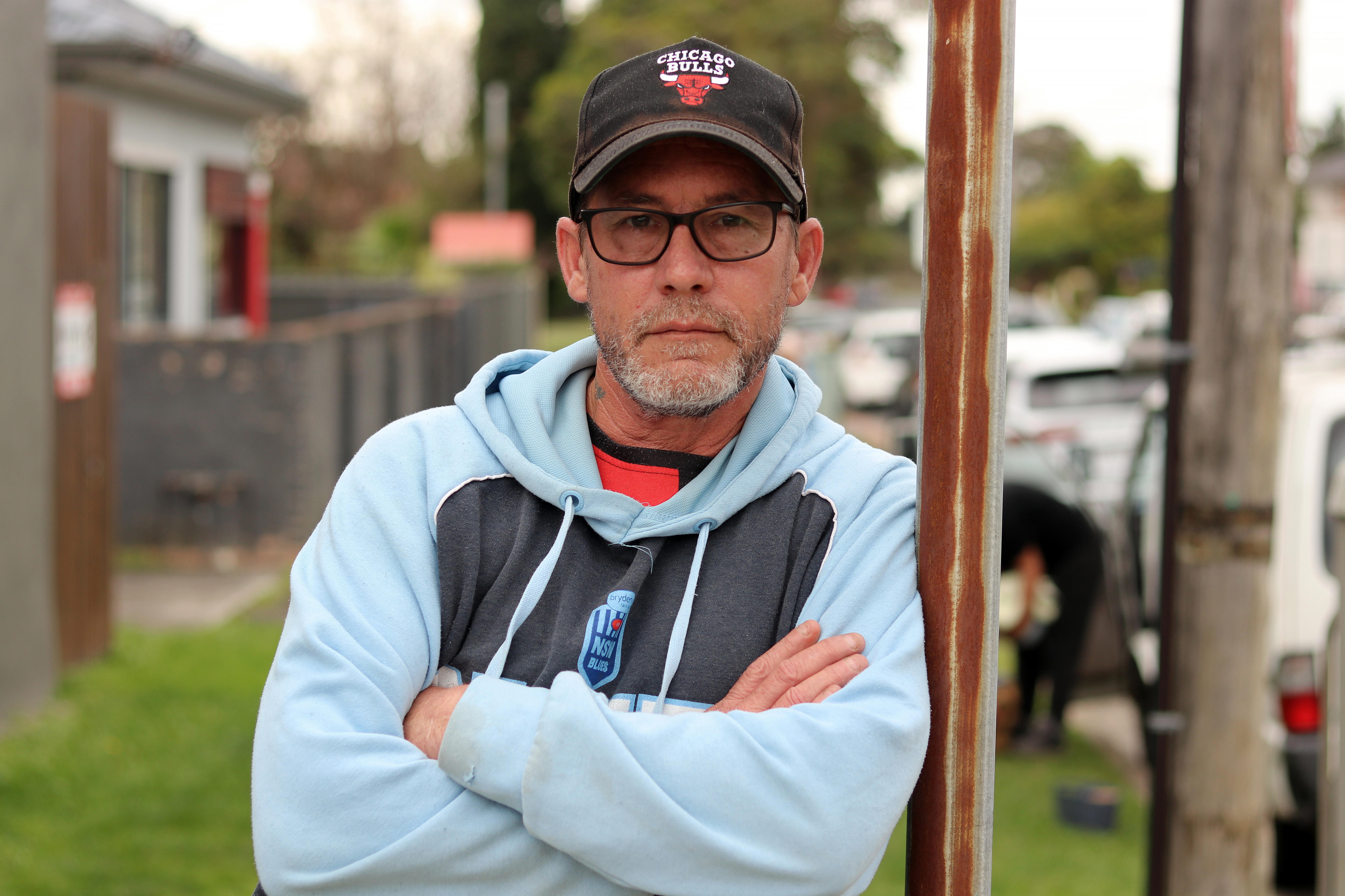 man in cap and glasses leans against pole with serious expression