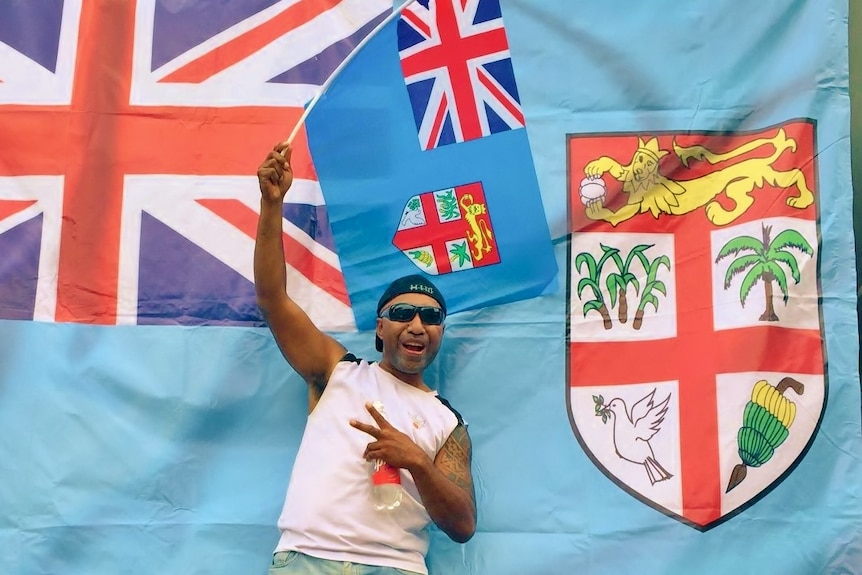 A Fijian man wearing sunglasses and a backwards cap waves a small Fijian flag while standing in front of a large one.