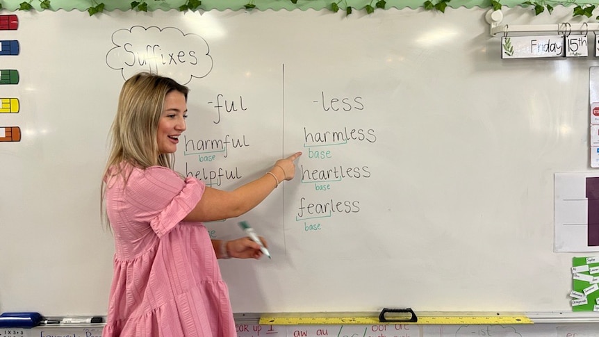 A smiling blonde woman teaches a class. She is making notes on a whiteboard.