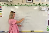 A smiling blonde woman teaches a class. She is making notes on a whiteboard.