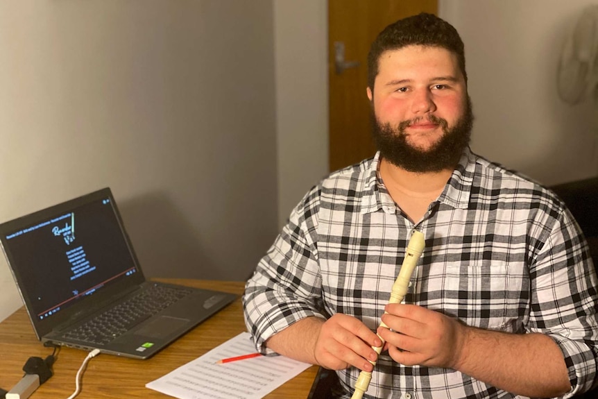 A bearded man holding a recorder while sitting at a desk with a sheet of music and a computer open.