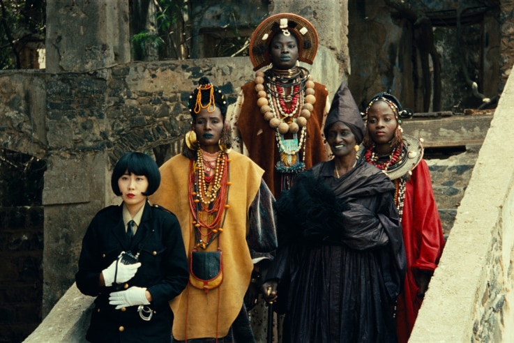 A still from 1992 film Hyenas with a group of African women in traditional dress and a Japanese woman in a military-style jacket