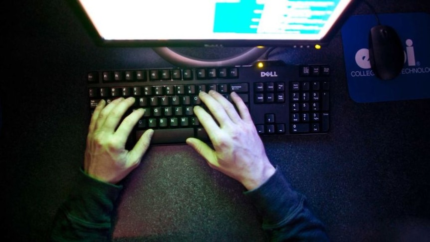 A person's hands are illuminated by the pale glow of a computer screen, as they type in a dark room.