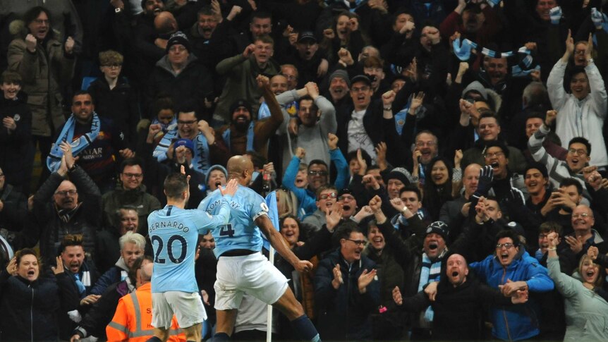 Vincent Kompany, wearing a light blue shirt, runs towards supporters who are all cheering