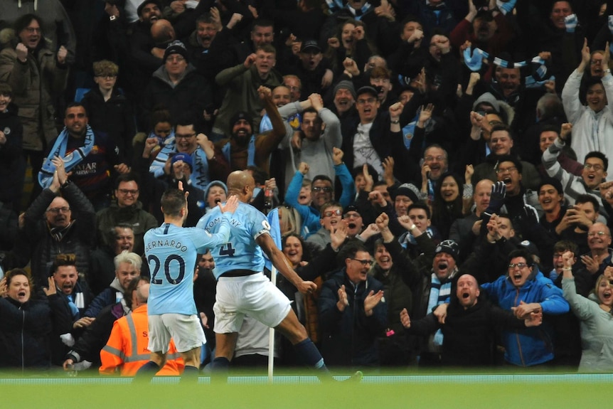 Vincent Kompany, wearing a light blue shirt, runs towards supporters who are all cheering