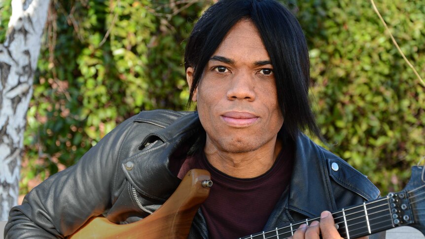 Stanley Jordan in a leather jacket, sitting in front of a hedge and staring at the camera as he plays the guitar