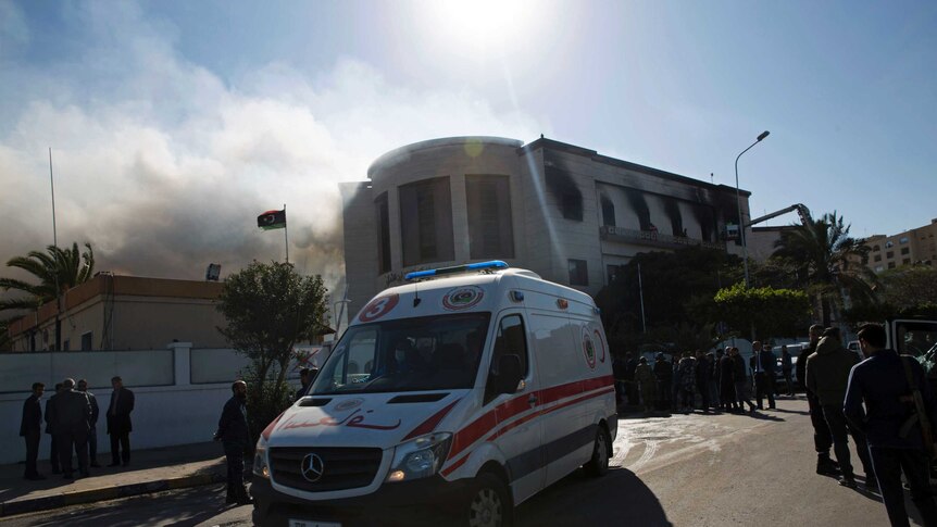 An ambulance waits outside of the burnt-out Libyan foreign ministry, with smoke billowing out to the left of frame.