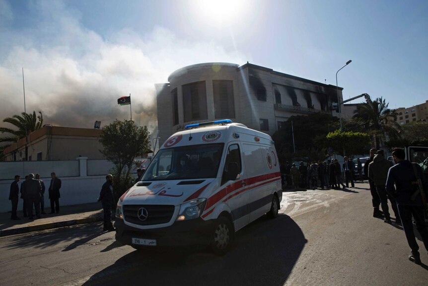 An ambulance waits outside of the burnt-out Libyan foreign ministry, with smoke billowing out to the left of frame.