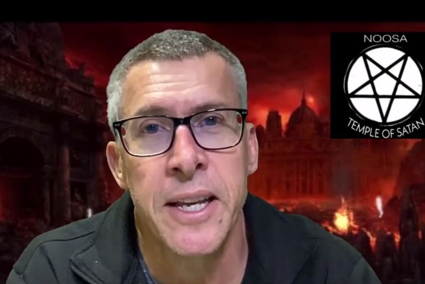 A man with gray hair wearing glasses sitting in front of a photo of a burning building, with Satan logo in corner