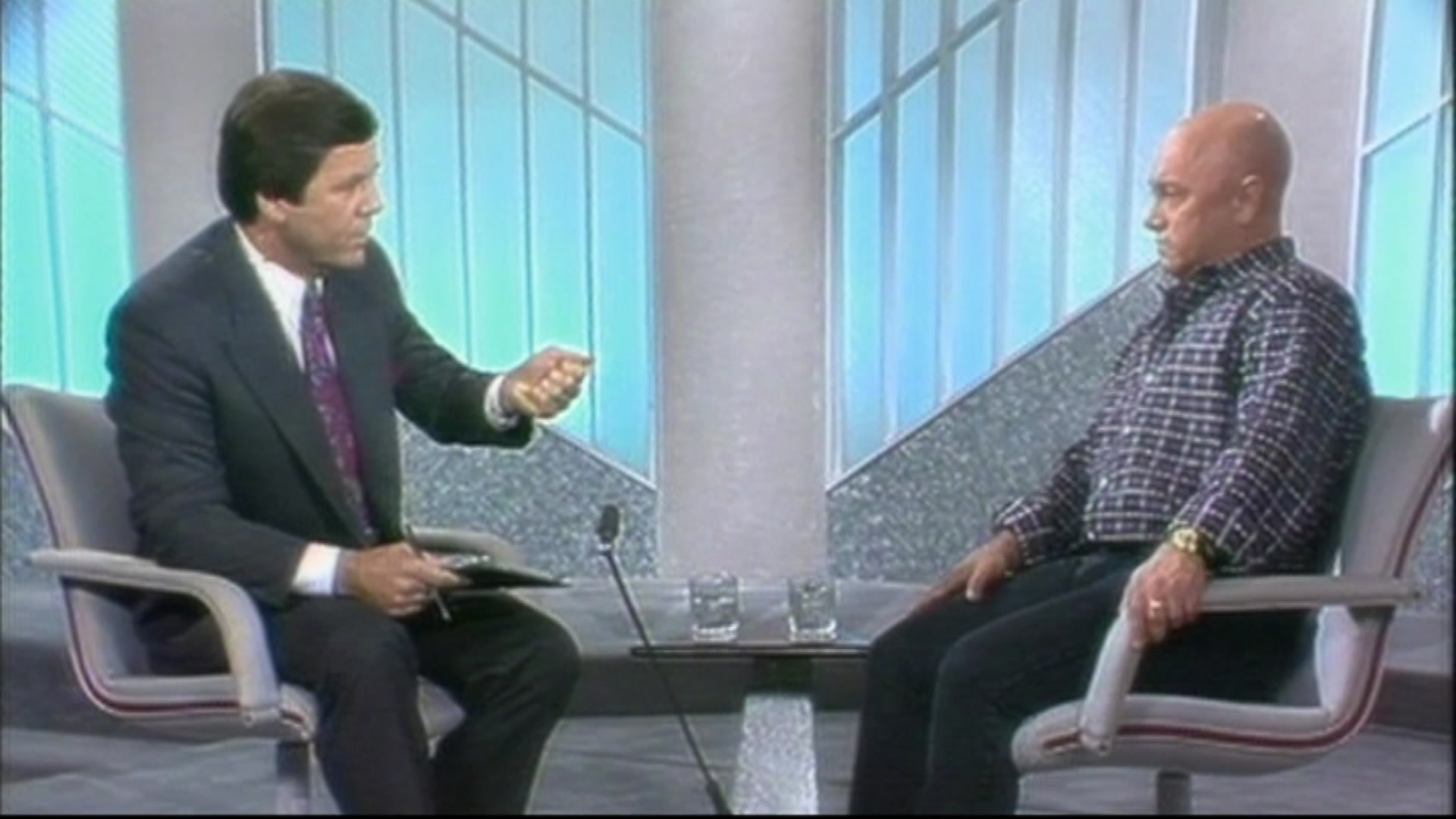 A dark haired man in a suit and a bald man in a checked shirt sit opposite each other in a tv studio.