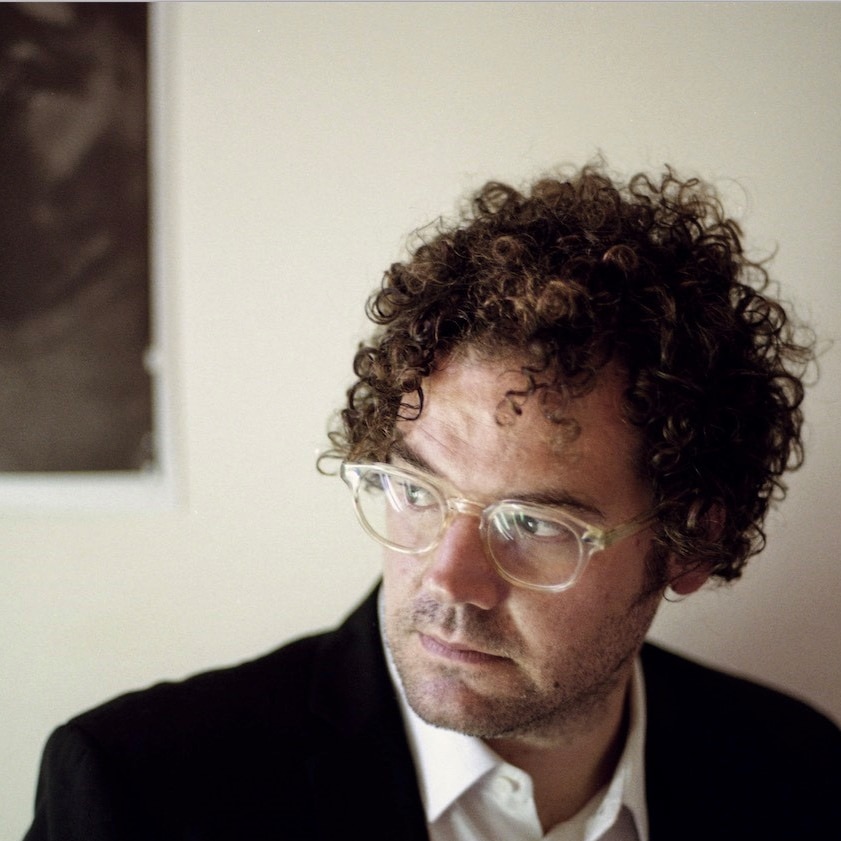 Laurence wears a white shirt and black suit jacket. He looks down to his right through clear-rimmed glasses.