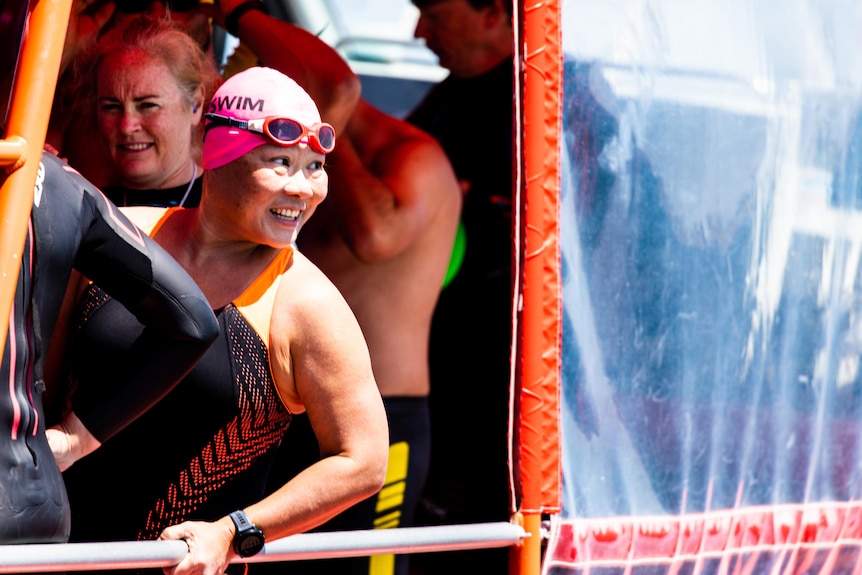 a woman in swimming gear on board a boat. she is looking out and smiling.