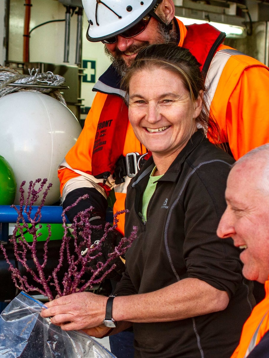 A woman smiles at the camera while holding up a piece of spiny, purple coral. Two men are smiling next to her.