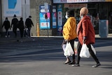 Two women holding shopping bags and wearing colourful puffer jackets and face masks walk across a road.