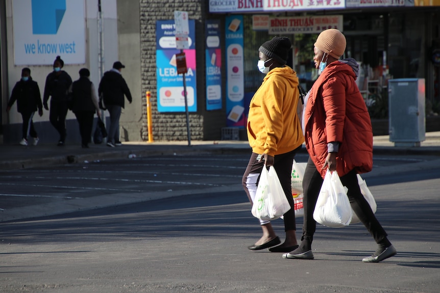 Deux femmes tenant des sacs à provisions et portant des doudounes colorées et des masques faciaux traversent une route.