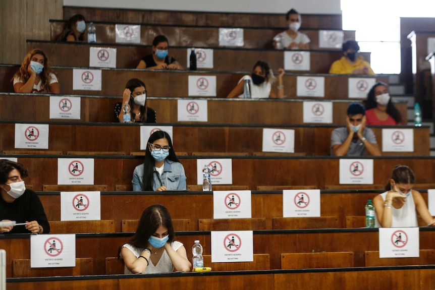 Students sit at a distance in rows of desks. Signs on every second seat block them from use.