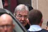 Prison guards surround George Pell as he leaves a prison van outside the stone walls of the court.