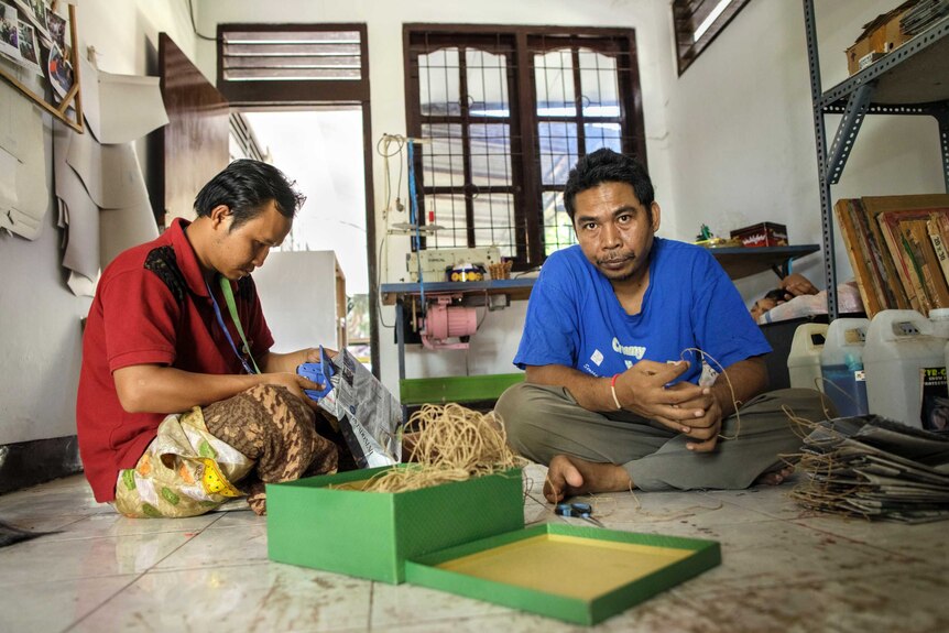 Two men sitting on the floor with one man making a paper bag and another man looking at camera