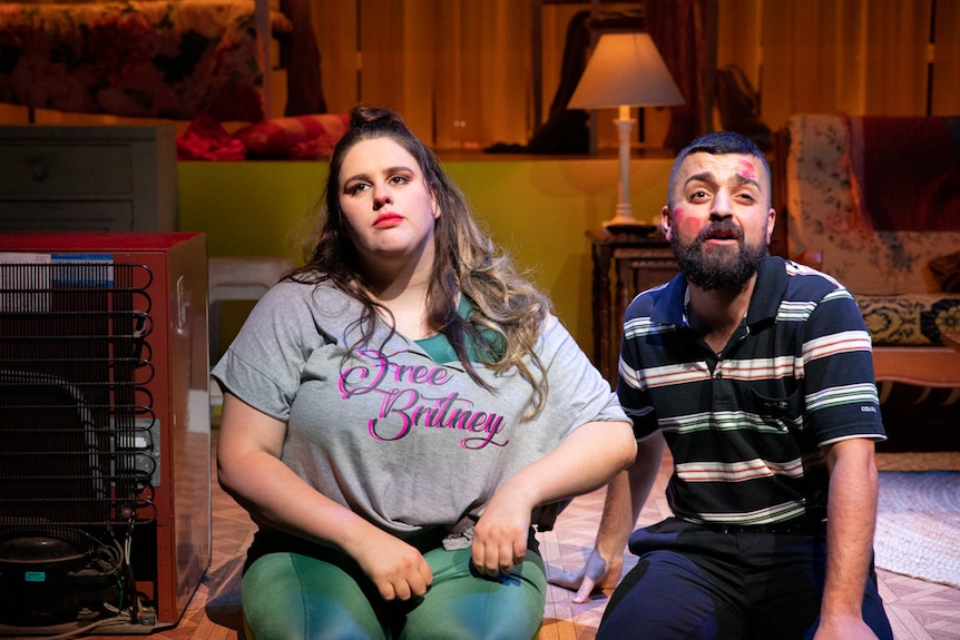 Interior living room; young woman in "Free Britney" tee shirt sits next to young man in polo shirt with lipstick kisses on face.