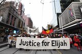 Protesters march down Bourke street in Melbourne with a banner that says 'Justice for Elijah'.