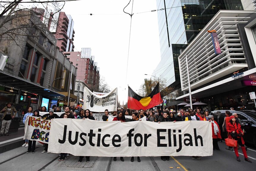 Protesters march down Bourke street in Melbourne with a banner that says 'Justice for Elijah'.