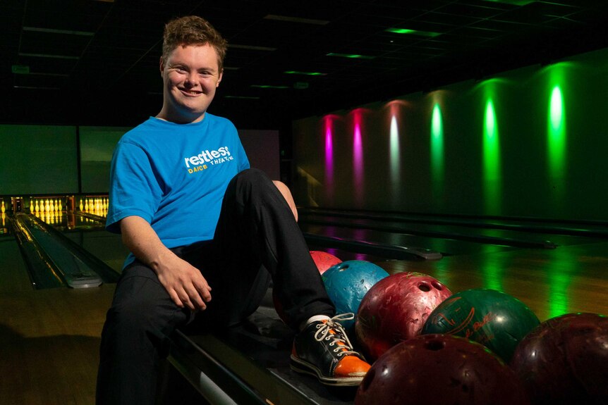A man sitting on a ledge in a darkened bowling alley, wearing a blue Restless Dance Theatre shirt.