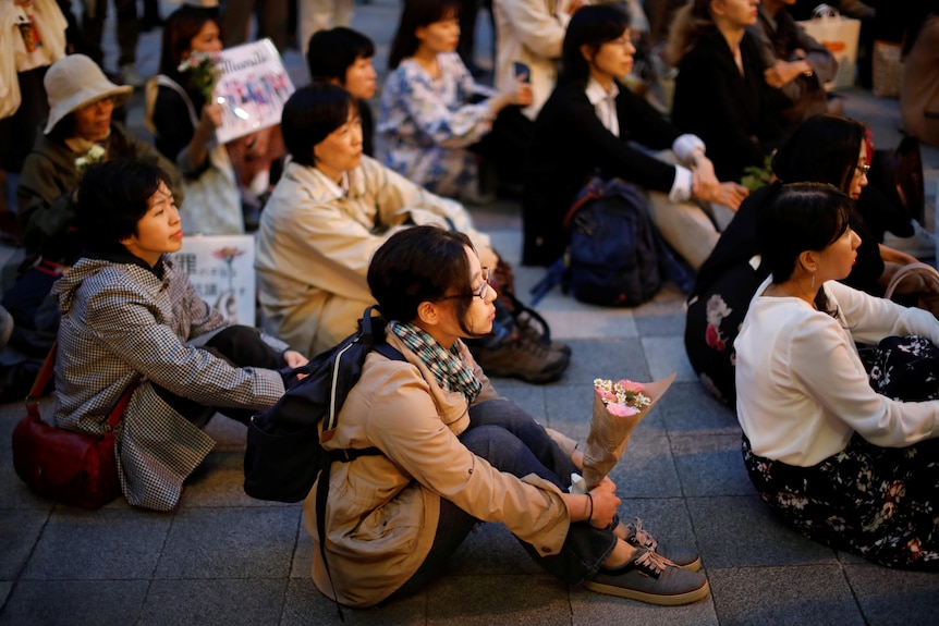 A group of women sit on the ground, some holding signs, some with candles