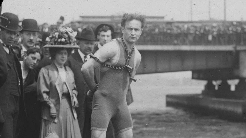A man in a swimsuit and chains stands on the top of a bridge in a 1900s photograph