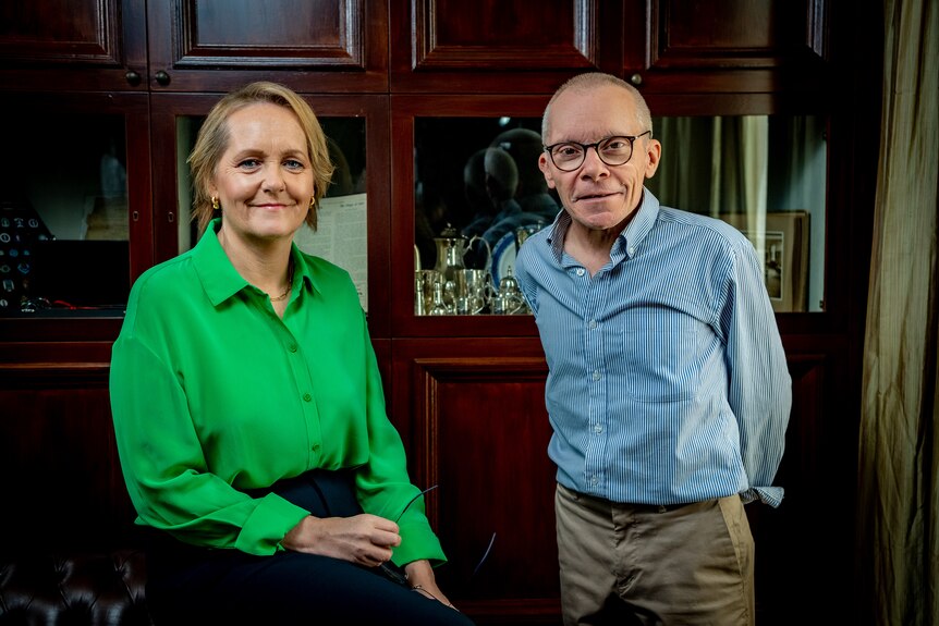 Woman with short blonde hair wearing a green top and man standing wearing a blue top and glasses.